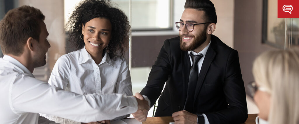 Two men in business attire are sitting at a table, shaking hands and smiling, with a woman beside them who is also smiling. They are likely discussing the new sales playbook in a professional meeting or interview setting, which exudes a bright and welcoming atmosphere.