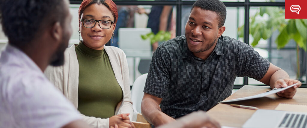 Three people sit at a table in lively discussion—a flipbook open beside them. One gestures while holding a laptop, another listens attentively, and the third smiles. The room is dotted with plants, enhancing the office setting's vibrant atmosphere.