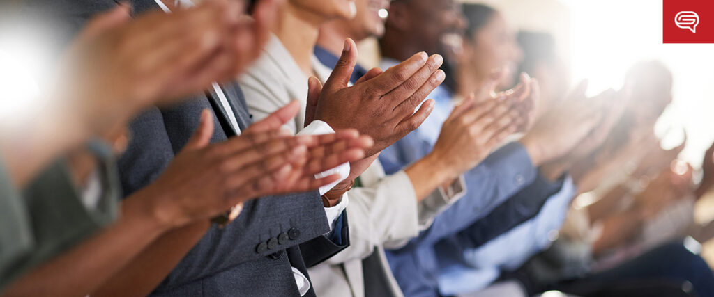 A diverse group of people sit in a row, their hands energetically clapping. In this formal setting, possibly a tech presentation, the focus on their hands highlights the visual elements that captivate the audience. Bright light streams in from the background, adding to the ambiance.