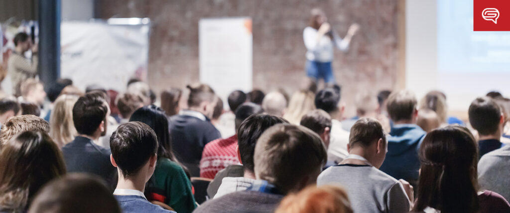 A large audience sits in a conference room, facing a speaker who stands in front of a screen displaying an interactive flipbook. The room is brightly lit, with a brick wall in the background and a logo in the top right corner.
