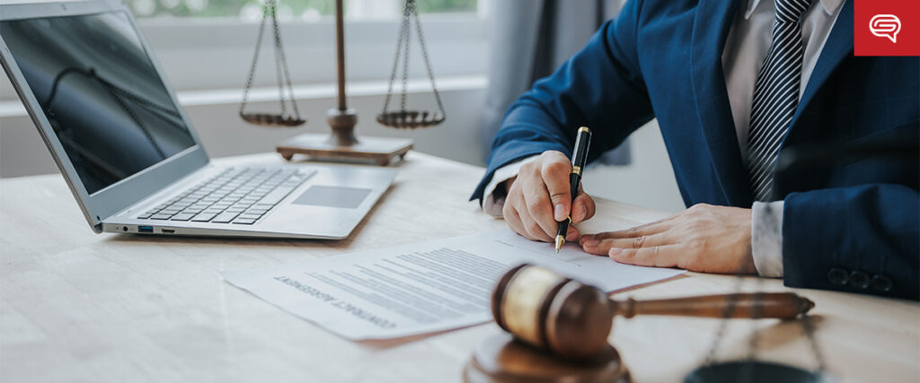 A person wearing a suit is signing a document on a desk. On the desk, there is a laptop displaying animations in pitch decks, alongside a gavel and a set of scales. The background shows a window with natural light coming in.
