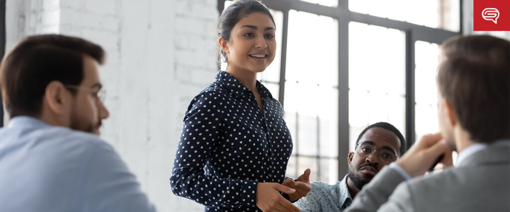 A woman stands and speaks to three colleagues seated around a table in a well-lit, modern office. She is smiling and wearing a navy blue polka dot blouse. One person is turned towards her attentively, while another appears engaged and thoughtful, likely discussing their latest quality earnings report.