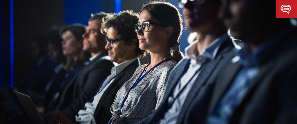 A group of people sits attentively in a dimly lit auditorium, all wearing business attire and lanyards. Most are focused straight ahead, possibly listening to a speaker or watching presentations on accessible healthcare. A woman in the center, wearing glasses, looks particularly engaged.