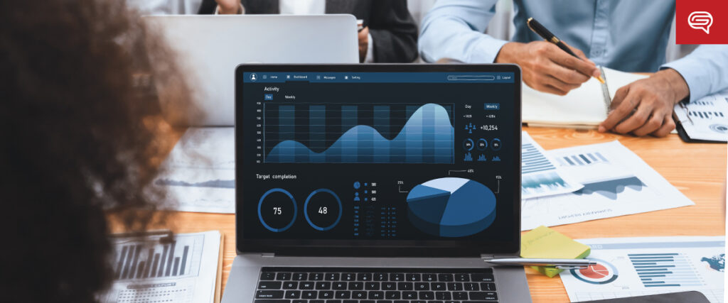 A laptop on a table displays various graphs and charts, showing blue-toned data visualizations. People sit around the table with documents and notes, some holding pens and writing. The atmosphere suggests a business meeting or data analysis session, possibly discussing accessible healthcare presentations.