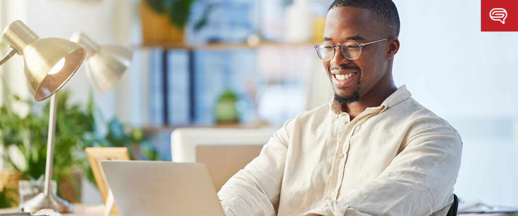 A man wearing glasses and a light-colored shirt is smiling and working on a laptop at a desk in a bright, modern office space. There is a lamp, some plants, and office supplies in the background as he refines animations in pitch decks.