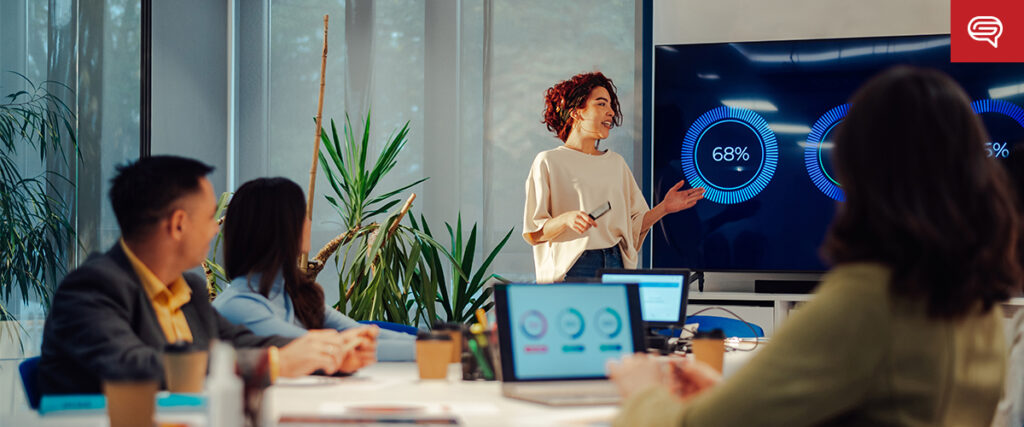 A woman is giving a quality presentation to three seated colleagues in a modern office. Large screens display charts showing percentages from the latest earnings report. The setting includes large windows and greenery, creating a bright, engaging workspace. Laptops are open on the desk.