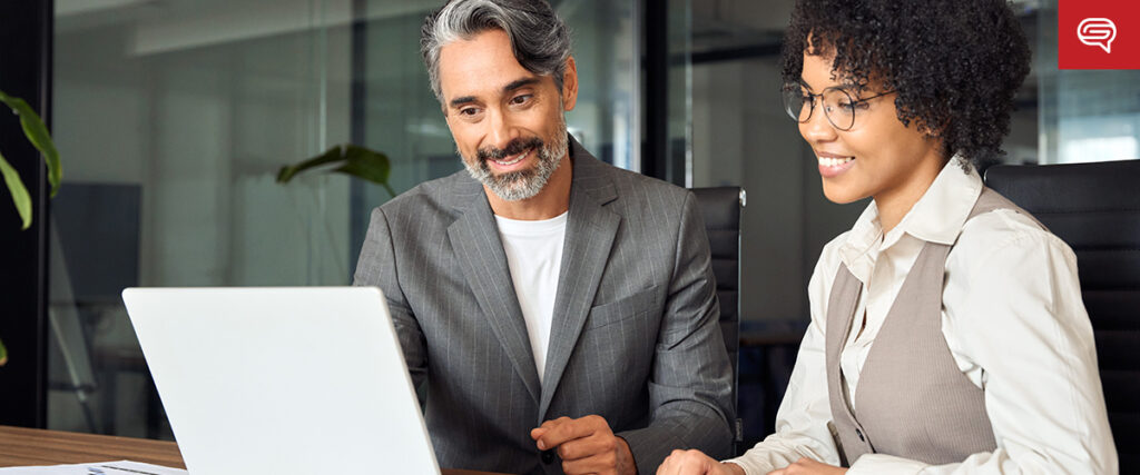 A man and a woman are sitting at a desk, both looking at a laptop and smiling. The man is wearing a gray suit while the woman is wearing glasses, a white shirt, and a beige vest. They appear to be in an office setting, engaged in preparing quality earnings report presentations.