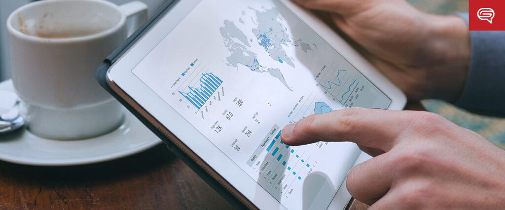 A person uses a tablet displaying various charts and a world map. The tablet shows graphical data, including bar charts and line graphs, as the individual interacts by pointing at one of the charts. Nearby on the table, a cup of coffee sits next to quality earnings report presentations.