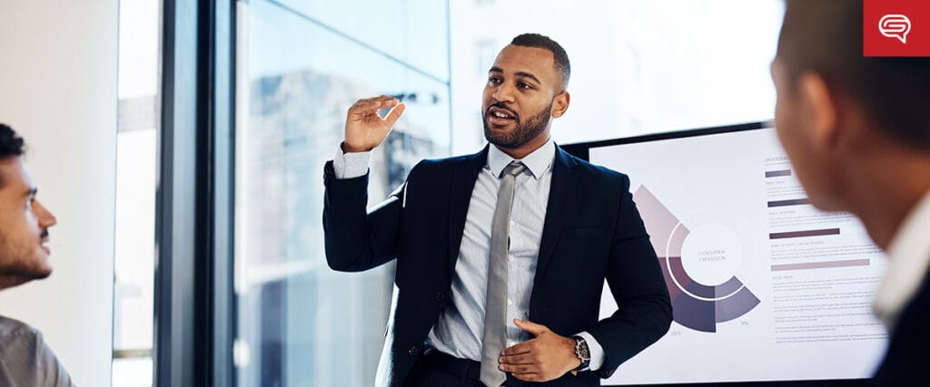 A man in a suit is giving a presentation in an office, skillfully using visual techniques to influence decisions. He gestures while addressing colleagues seated around a table, as a screen displays case study slides with charts and text. Bright windows and modern furnishings are visible in the background.