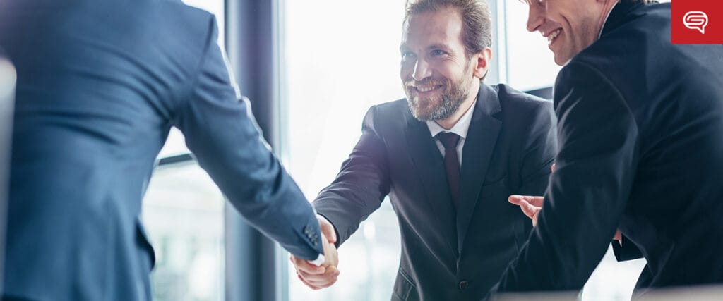 Three men in suits engage in a handshake in a well-lit office, all smiles and positivity suggesting a successful business meeting. A red logo is prominently displayed, perhaps indicating a fruitful discussion about consumer electronics or an important patent.
