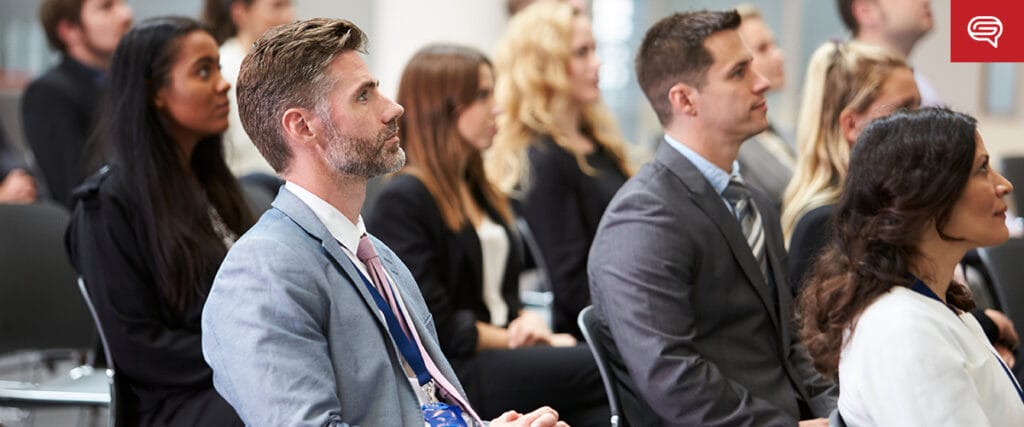 A diverse group of people seated in a conference room, attentively listening to a presentation on case study formats. Dressed in formal attire, with notebooks and lanyards visible, they engage in this professional seminar designed to enhance visual communication skills.