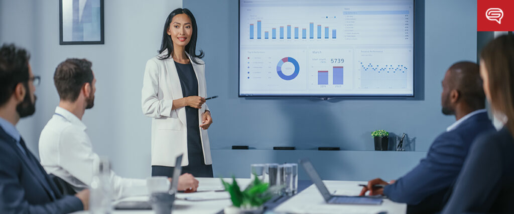 A woman presents to a group in a conference room, gesturing towards a slide displaying charts and data on consumer electronics. The audience, seated at a table with laptops, listens attentively. The setting is professional and business-oriented.