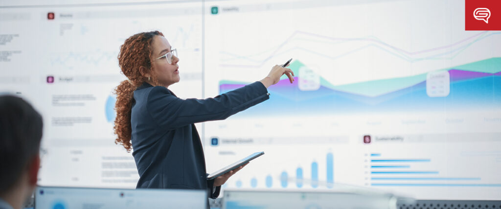 A woman in a business suit points at a large digital screen displaying graphs and charts, seamlessly blending visual techniques with her presentation. Holding a tablet, she explains data that influence decisions in the modern office where attentive people listen to her insights.
