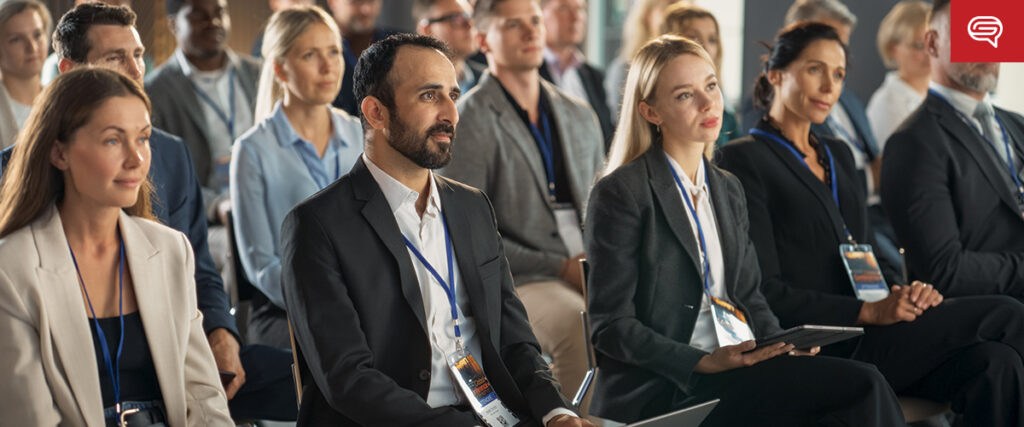 A diverse group of people sits attentively at a conference, ready to influence decisions. Dressed in business attire and wearing name badges, they eagerly follow along with notebooks and devices in hand, as case study slides illuminate the room.