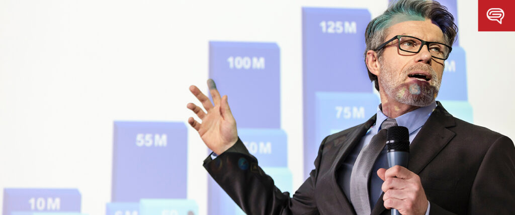 A man in a suit and glasses speaks into a microphone at an investor presentation, gesturing at a screen displaying a bar graph with figures ranging from 10M to 125M. The background is white with a red logo in the top right corner.