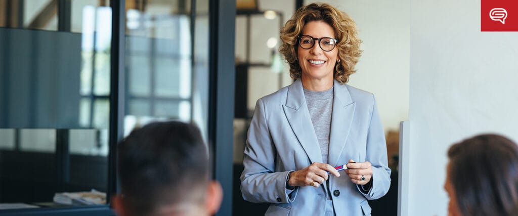 A person with curly hair and glasses stands smiling in an office, holding a marker. They are facing two seated individuals, engaging in a lively presentation on seed funding round strategies. The room features glass partitions and modern decor.