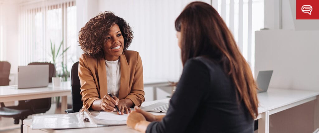 Two women sit at a desk in an office with laptops, preparing for seed funding round presentations. One woman with curly hair in a tan blazer is smiling and holding a pen, while the other has long brown hair. Both appear engaged in conversation. A red logo is in the top right corner.