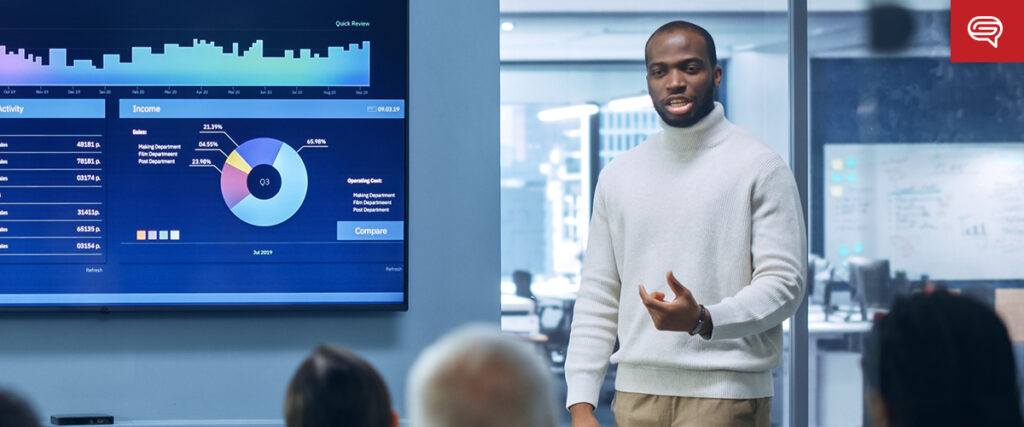 A person is leading a seed funding round presentation in an office, standing next to a screen displaying charts and graphs. The presenter, in a light-colored sweater, engages the audience seated before them. The room is modern with glass walls and office equipment.