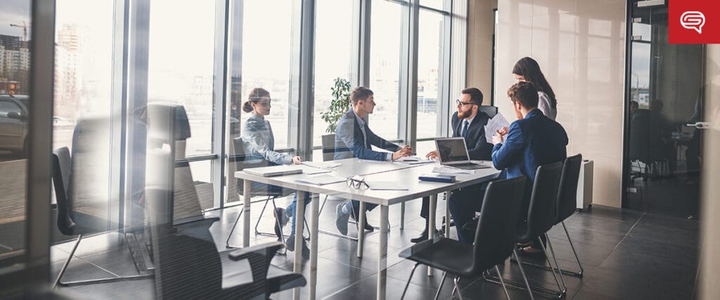 Five people in a modern office meeting room are engaged in discussions around the table, laptops and documents scattered, as they refine their seed funding round presentations. Large windows flood the space with natural light, while a plant adds a touch of greenery to the focused atmosphere.