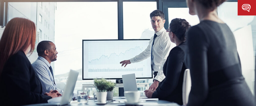 A man in a white shirt presents a line graph during a seed funding round presentation to four colleagues. They are seated around a table with laptops and documents, while the room's large windows let in ample light, creating an atmosphere that appears both professional and engaged.