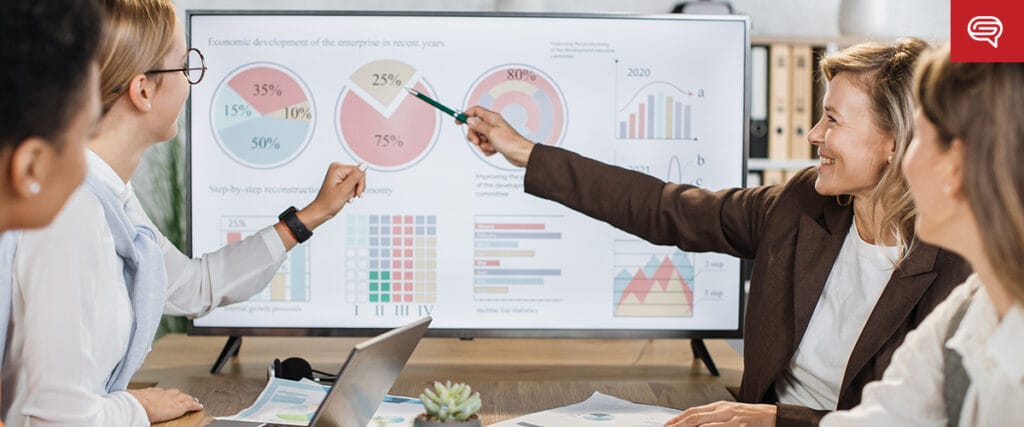 Four people are in a meeting, examining a presentation screen displaying charts and graphs about economic development. During this seed funding round, one person is pointing at the screen with a pen, passionately discussing the data.
