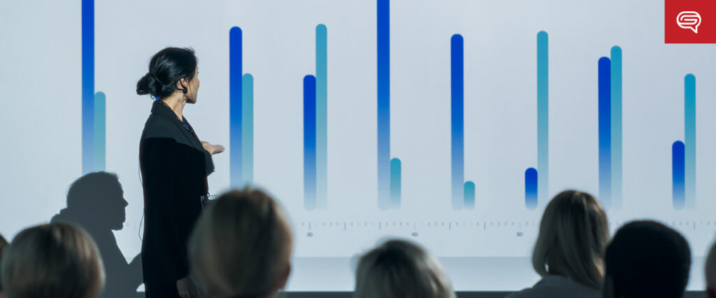 A woman in a dark blazer delivers a seed funding round presentation, showcasing a bar graph to an audience in a dimly lit room. The graph features blue vertical bars of varying heights. The audience is seated in the foreground, attentively facing the presenter and the screen.