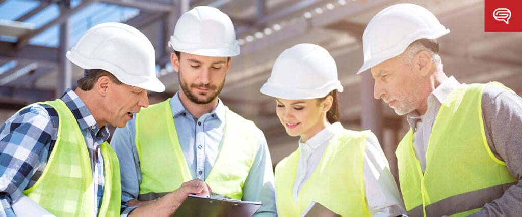 A group of four construction workers in hard hats and reflective vests stand at a construction site, reviewing documents on clipboards, much like executives fine-tuning their presentations before a crucial seed funding round. The building framework rises against the clear sky in the background.