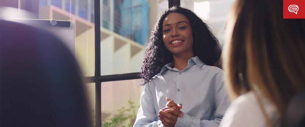 A woman with long curly hair smiles while standing in front of a group during seed funding round presentations. She is wearing a light blue shirt and clasping her hands. In the background, large windows let sunlight stream in, adding warmth to the room.