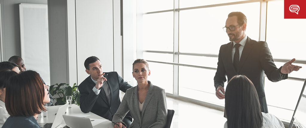 A group of people in business attire are gathered in a conference room. A man is standing and presenting near a large window, discussing seed funding round presentations, while others sit around a table, listening attentively.