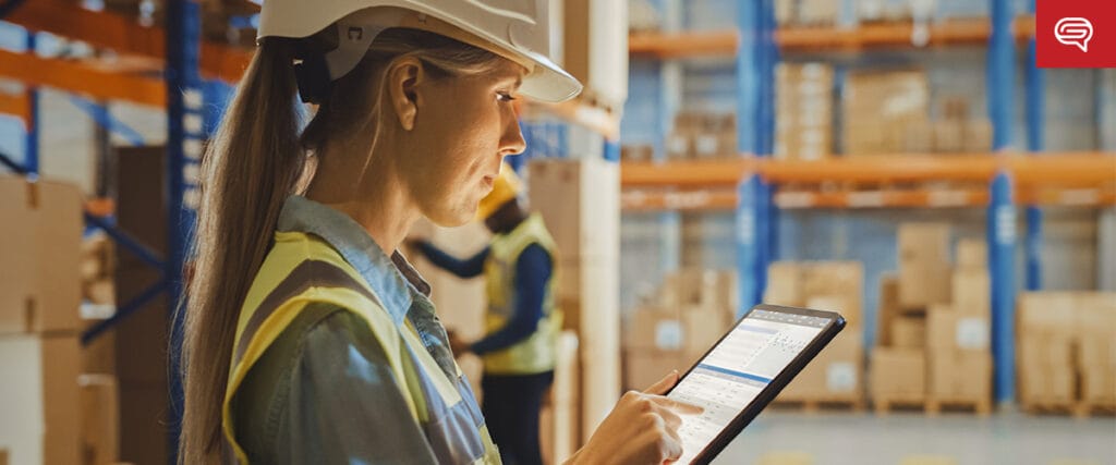 A woman in a hard hat and safety vest uses a tablet in a warehouse, enhancing client experience. Shelves stacked with boxes are visible in the background, while another worker inspects inventory, suggesting logistics or supply chain management akin to consulting firm operations.