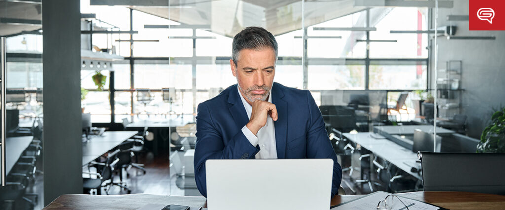 A man in a suit sits at a desk in a modern office, focused on his laptop, crafting a client experience slide for consulting firms. Natural light streams in through large windows, illuminating the room's sleek decor.