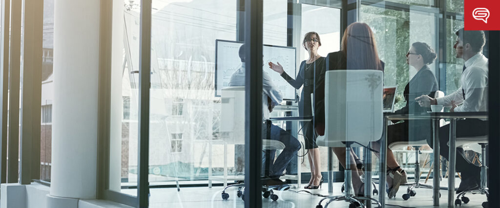 A group of people in business attire gathers in a modern glass-walled conference room. One person is presenting near a digital screen displaying charts on client experience, pivotal for consulting firms. Sunlight filters through large windows, illuminating the room.