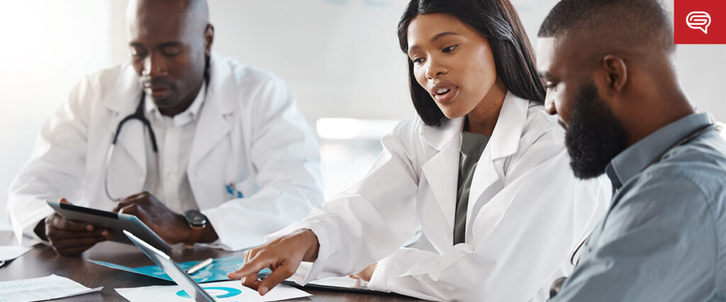 Three medical professionals in white coats sit around a table. One woman points at a document on a laptop while the man on her right looks on. Another man to her left holds a tablet, examining a client experience slide, as if preparing insights for consulting firms. They appear to be in a meeting.
