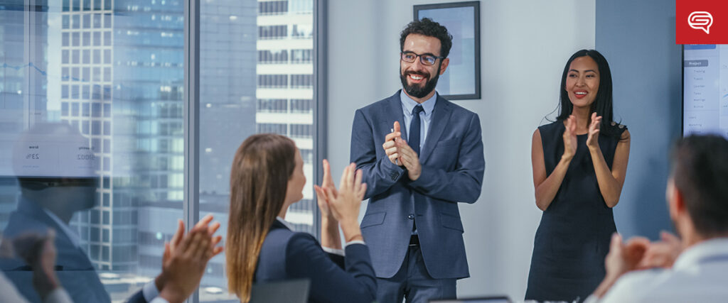In an office setting, a group of professionally dressed individuals applauds as a man in a suit and a woman in a dress stand at the front, presenting a client experience slide for consulting firms. The large windows behind them reveal towering city buildings.