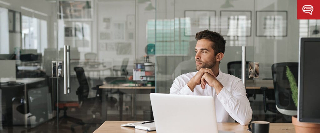 A man in a white shirt sits at a desk with a laptop in a modern office, reflecting on enhancing client experience slides for consulting firms. Gazing off to the side, he rests his chin on his hands. The softly lit office features glass walls and chairs scattered among desks in the background.