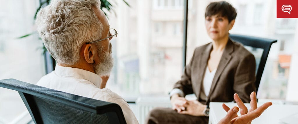 A man with gray hair, seated with his back to the camera, gestures while discussing client experience slides with a woman in an office setting. She is wearing a brown suit and listening attentively. Large windows in the background offer a view of buildings outside.
