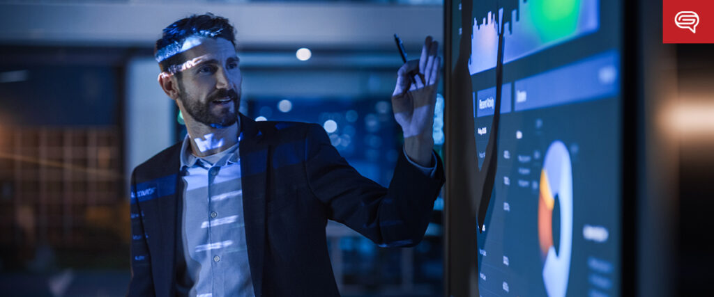 A man in a suit stands before a large screen displaying graphs and charts, pointing at data with a pen. This client experience slide, crucial for consulting firms, is highlighted as the dimly lit room emphasizes the illuminated visuals.