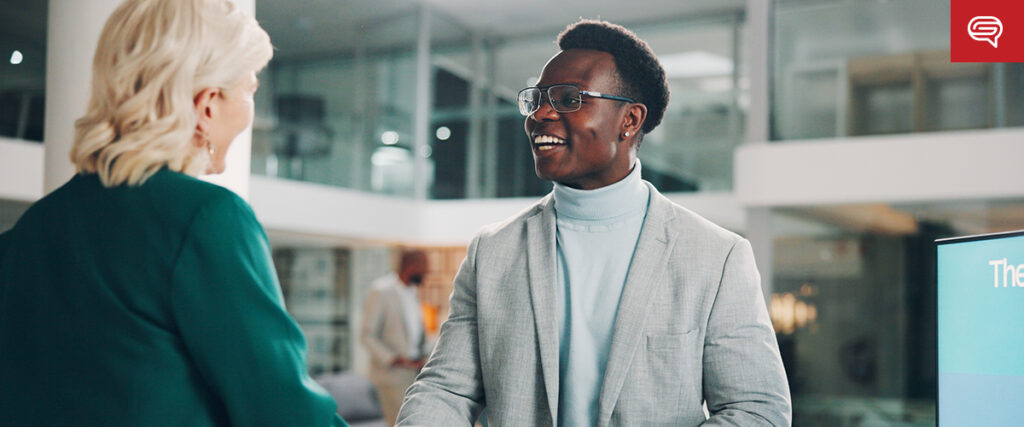 A man in a light suit and glasses shares a warm handshake with a woman in a green outfit, enhancing the client experience. They stand in a modern office with large windows and a banner, resembling an ideal slide for consulting firms.