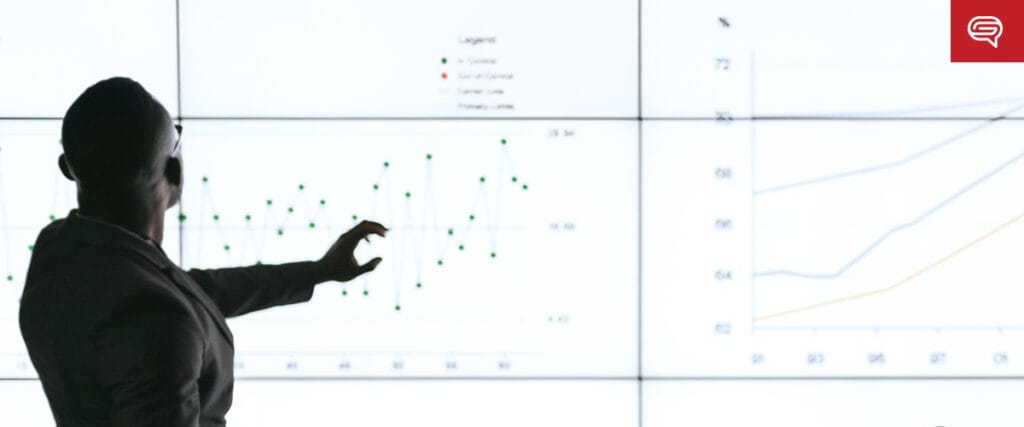 A person in a suit points to graphs on a large screen, highlighting client experience trends. The vibrant display offers insights tailored for consulting firms, casting the presenter in silhouette against the bright background.