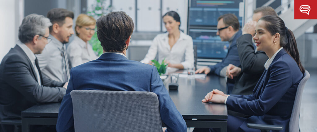A group of business professionals in formal attire sit around a long table in a modern meeting room, where visual presentations on large screens boost engagement. The atmosphere appears focused, enhancing their discussion on real estate sales.