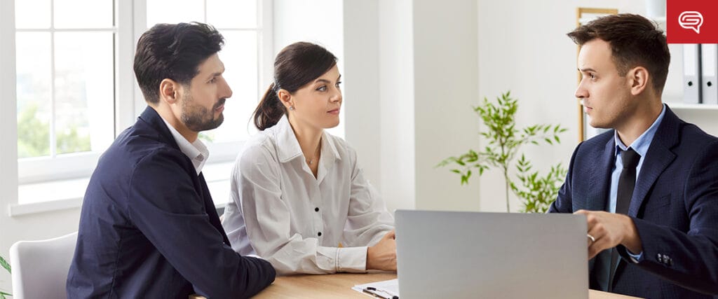 A couple sits across a table from a man in a suit in a bright office, discussing their startup's branding strategy. The man speaks animatedly, gesturing towards the laptop. The couple looks attentive and engaged, surrounded by large windows and lush plants in the background.