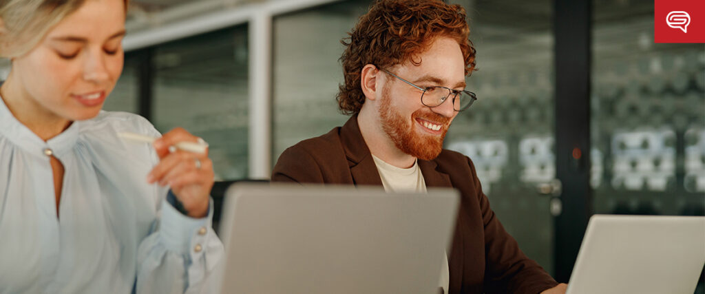 A man with red hair and glasses is smiling while using a laptop, engaged in hiring a presentation design agency. A woman beside him, partially visible, appears focused on her laptop. They are in a modern office environment with a black and glass wall in the background.