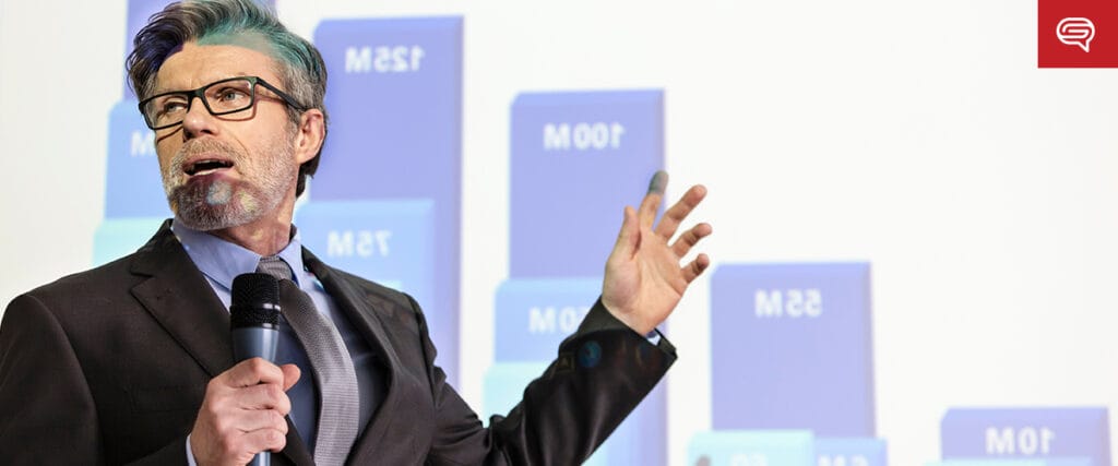A man in a suit and tie is speaking passionately about branding strategy while gesturing towards a large bar graph projected on a screen. The graph displays various numbers, such as 100M and 25M. He seems to be giving a presentation related to consumer goods.