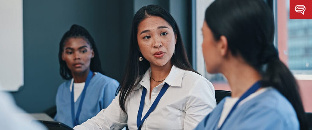 Three women, two in blue scrubs and one in a white shirt, sit together in a meeting room. The woman in the center speaks while the others listen attentively, with a red speech bubble icon and a market share slide for tech investors visible.