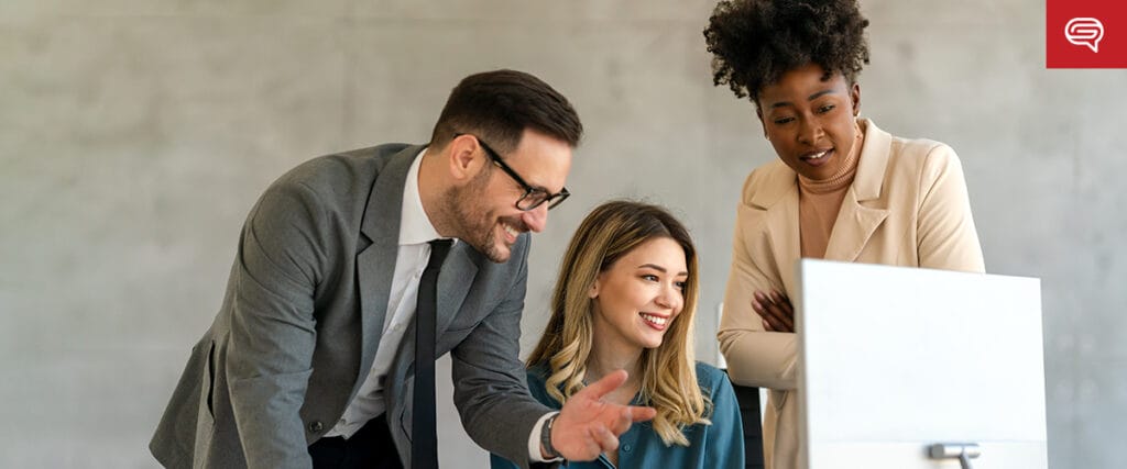 Three people in business attire gathered around a computer screen, looking engaged. A man on the left gestures towards the branding strategy slide, while two women smile, observing intently. They are in an office setting with a gray background.