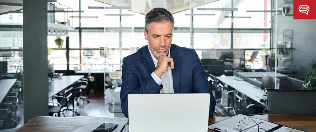 A man in a blue suit sits at a desk in an office, thoughtfully preparing a visual presentation on his laptop. Large windows and modern furniture frame the scene, while scattered papers hint at real estate sales strategies he plans to boost.