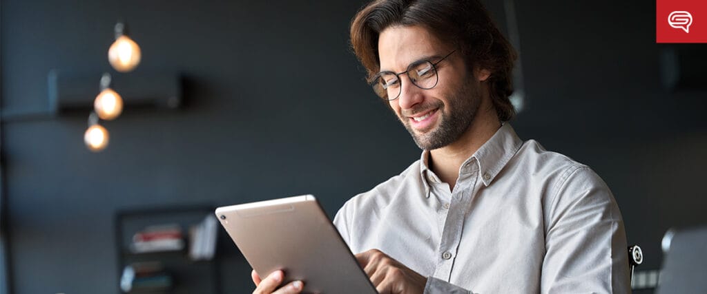 A man with glasses and a beard smiles while using a tablet in a modern, softly lit room—perfect for reviewing an executive summary slide. Spherical hanging lights and blurred shelves create an inviting background.