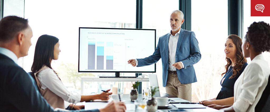 A man in business attire presents stacked bar graphs on a screen to four colleagues seated around a conference table in a modern office with large windows.
