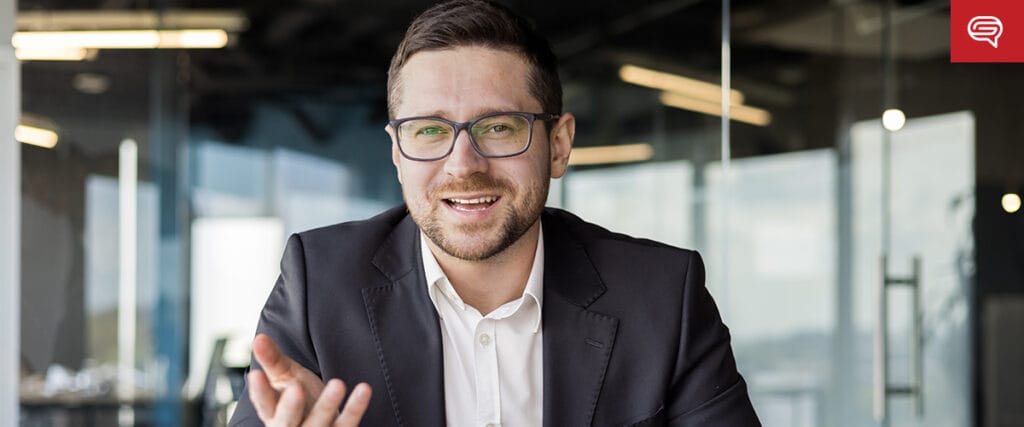 A man in a suit and glasses sits at a desk in a modern office, gesturing with one hand and smiling, as if presenting an executive summary slide during a conversation.