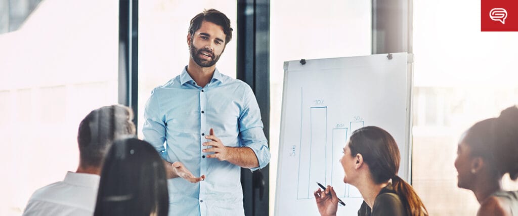A man stands and presents in front of a group, gesturing beside a flip chart showing bar graphs and geographic heatmaps, while seated colleagues listen and take notes in a modern office setting.